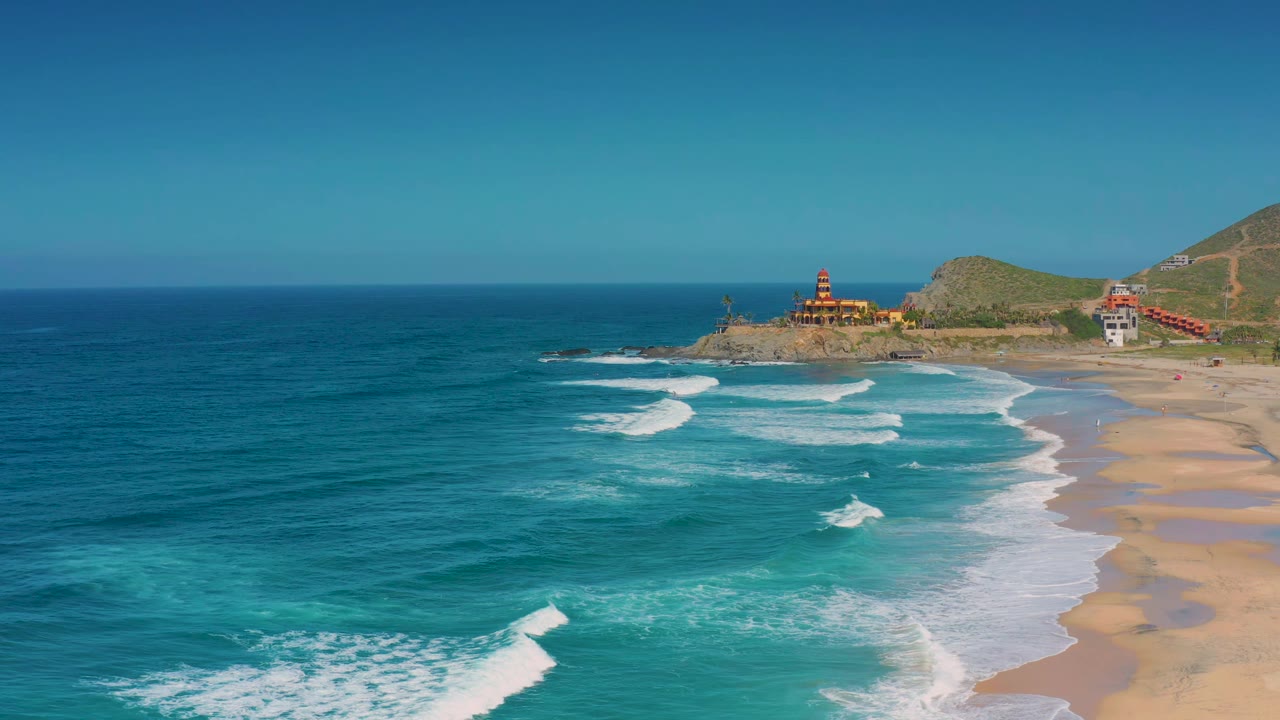 Wide drone view of surf lines curving toward a sandy shoreline and cliffs.