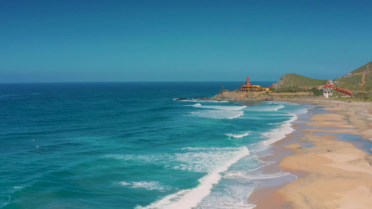 Wide aerial view of Cerritos, Baja California Sur, Mexico, where surf lines approach pale sand, deep blue water fills the left side, background hills rise behind coastal structures, and no visible people appear.