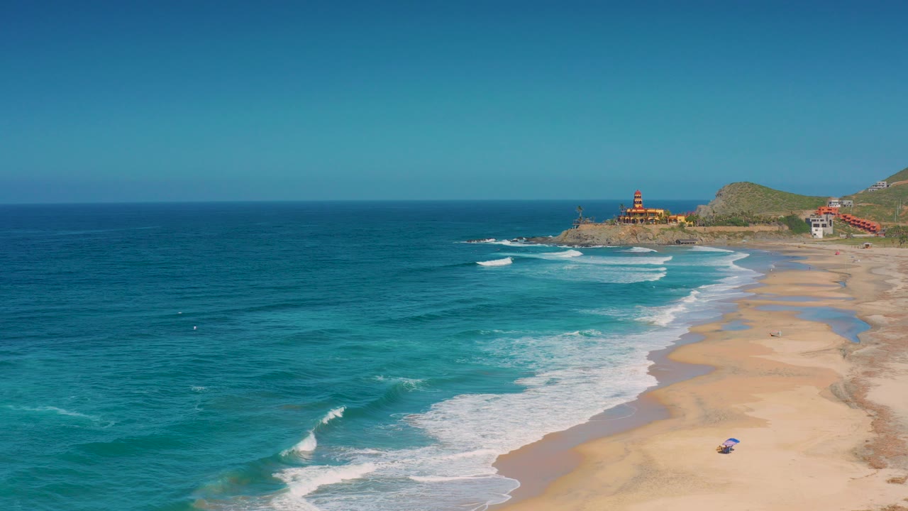 Wide aerial view over Cerritos, Baja California Sur, with blue ocean at left, white surf on a broad sandy beach, distant coastal buildings and no visible people.