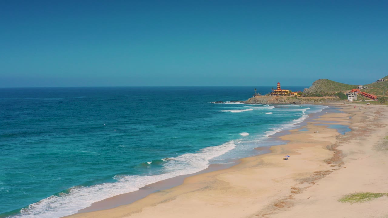 Wide aerial frame over Cerritos, Baja California Sur with turquoise water, white surf, a long sandy beach, distant headland buildings, and no visible people.