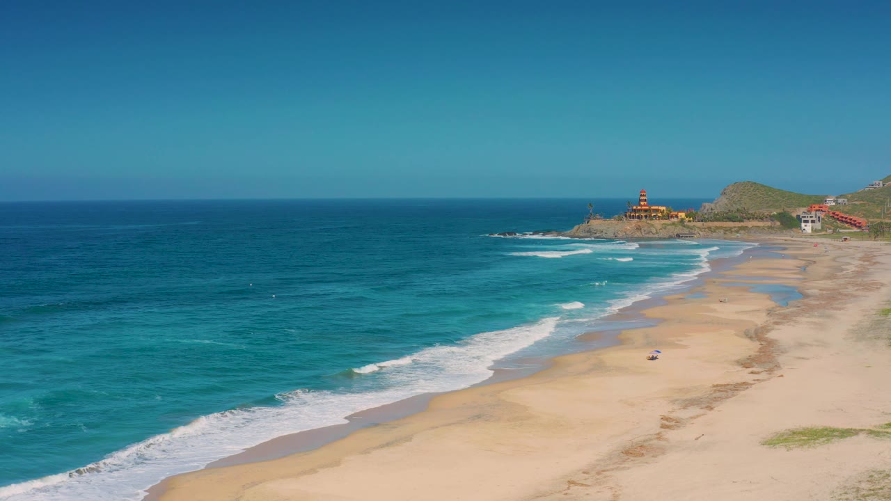 Aerial frame over Cerritos showing a wider sweep of water, foamy surf approaching the beach, tan sand, a far headland with structures, and no visible people.