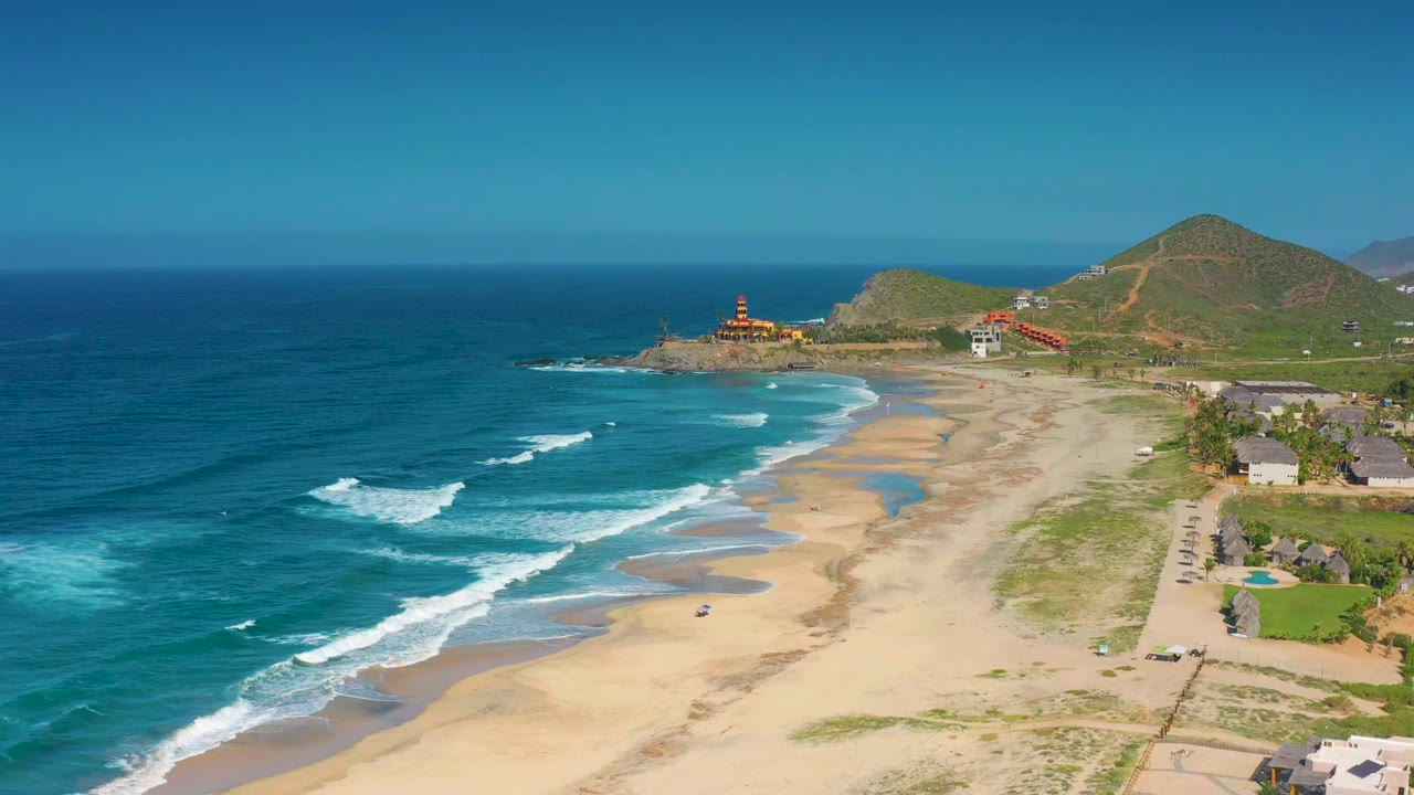 Aerial view over Cerritos, Baja California Sur, with blue ocean on the left, waves crossing the sand, low hills in the distance, scattered beachside buildings to the right, and no visible people.