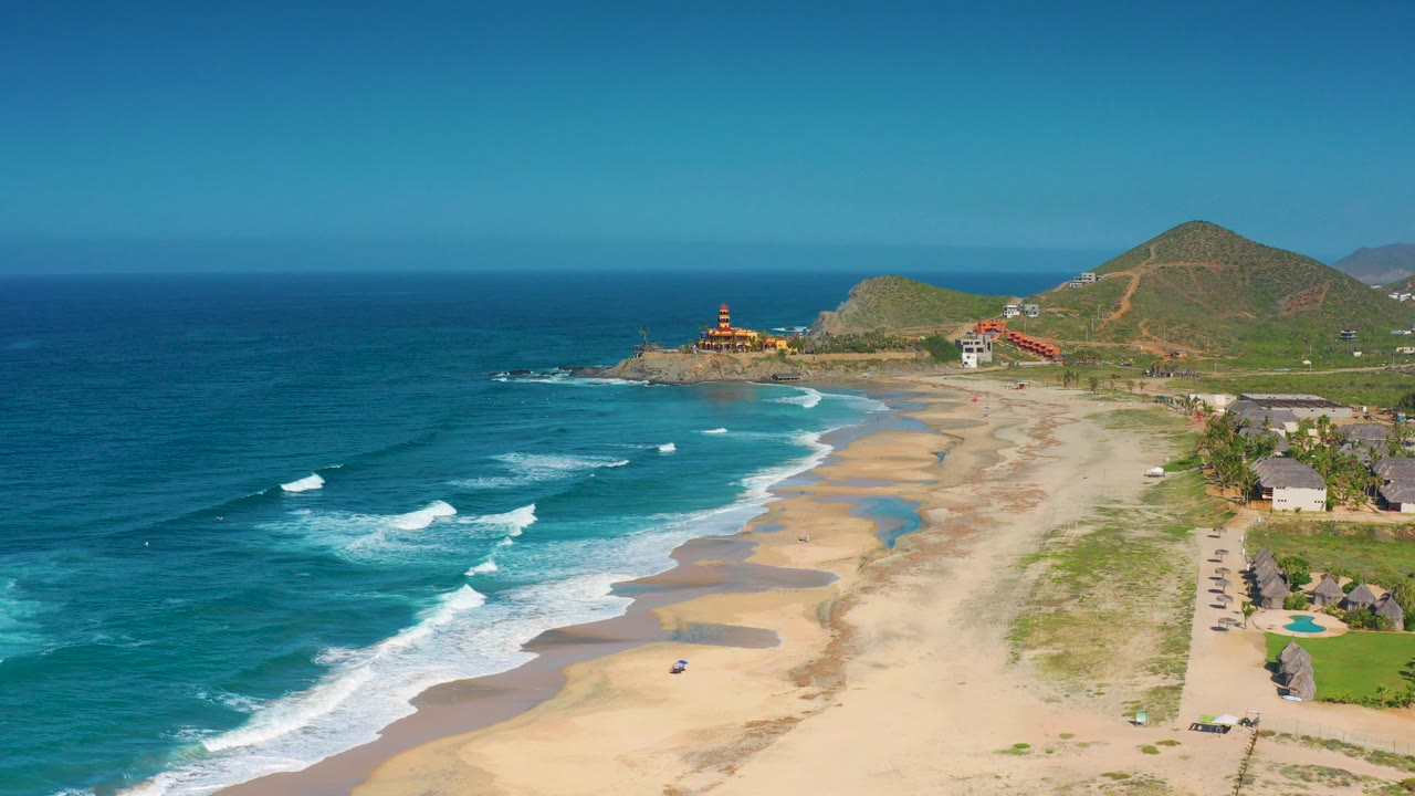 Opening aerial frame over Cerritos, Baja California Sur, showing blue ocean on the left, pale sand below, small beach houses on the right, green hills behind, a lighthouse on the point, and no visible people.