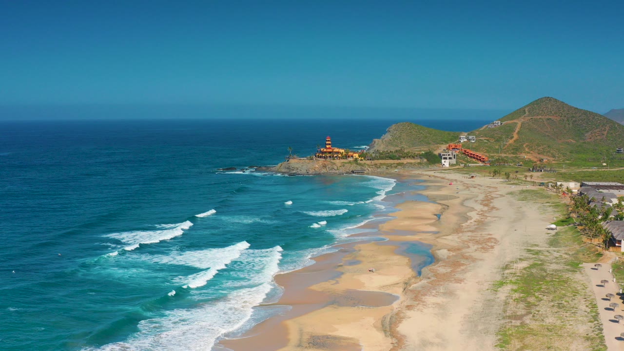 Aerial frame from Cerritos, Baja California Sur showing blue ocean at left, white surf lines reaching a wide sandy beach, low hills and distant buildings at right, with no visible people.