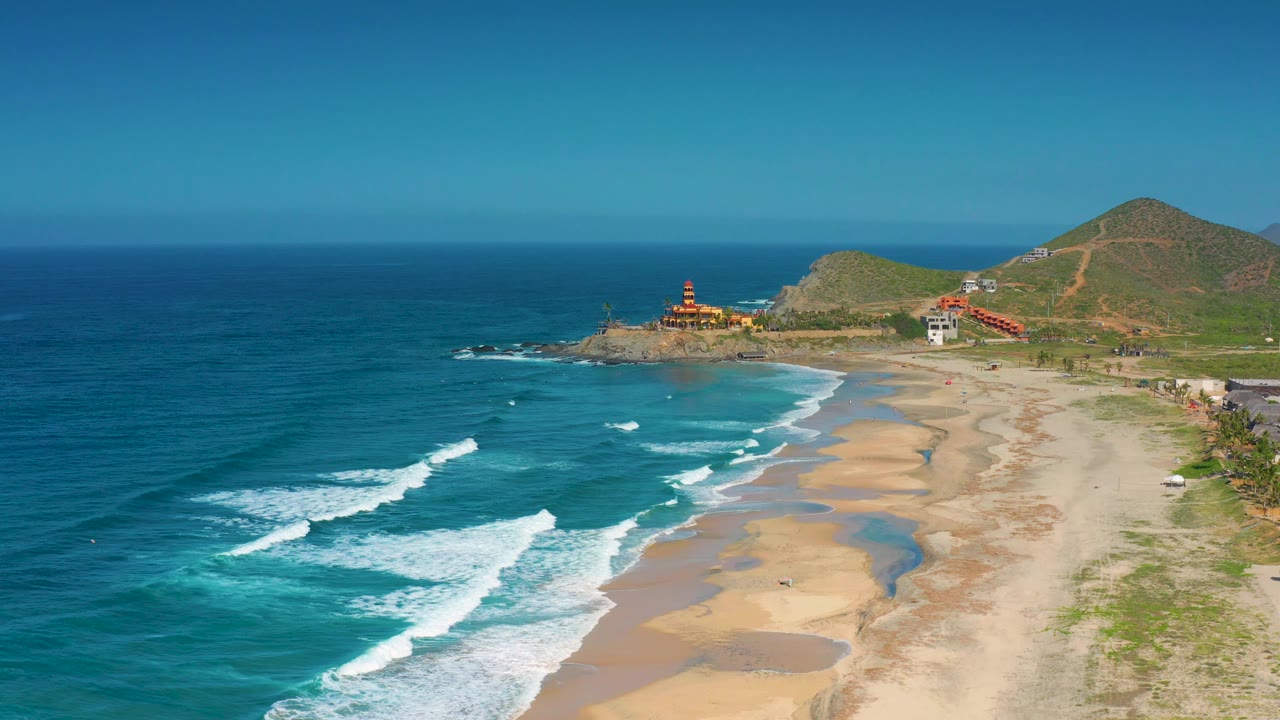 High coastal view of waves, beach, a rocky point, and hillside structures under clear sky.