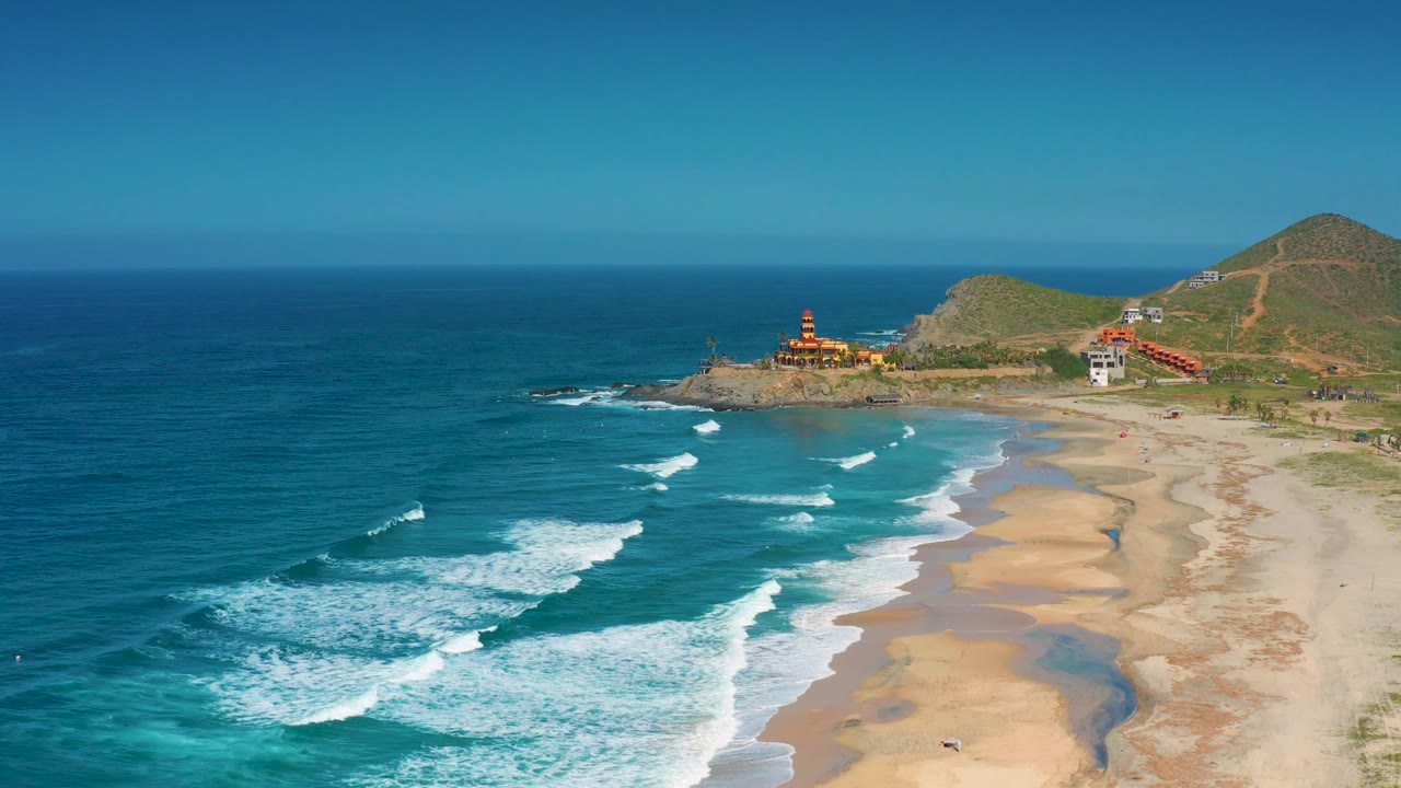 Wide Cerritos coastline view with foamy surf spread across blue-green water, broad sandy beach, rocky headland buildings, hills, and no visible people.