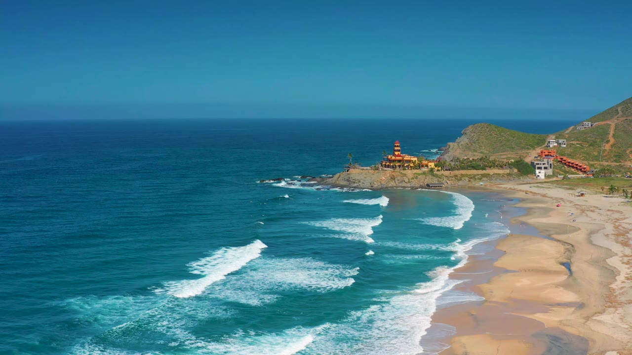 At 0:01, the Cerritos coastline remains wide in frame as parallel white waves approach tan sand, with the tower on the rocky point and no visible people on the beach.