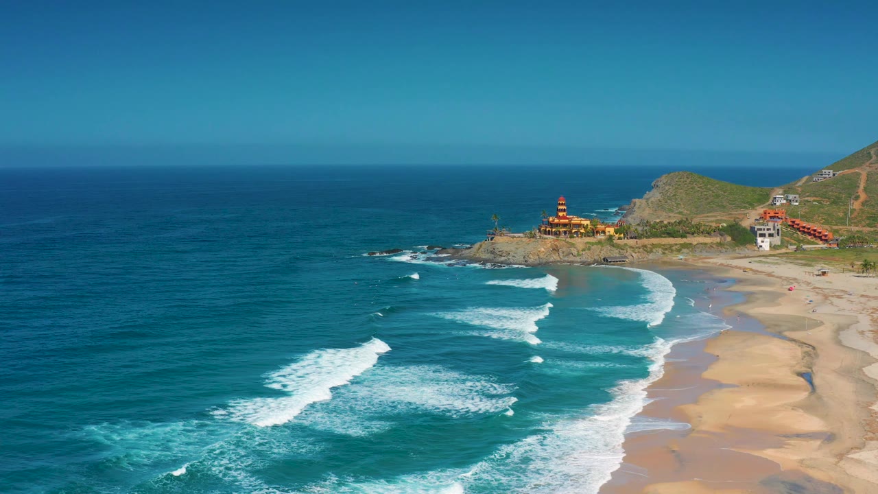At 0:02, the aerial view shows more of the sandy right edge while whitewater breaks across the center water, the small tower sits on the point, and no visible people appear.