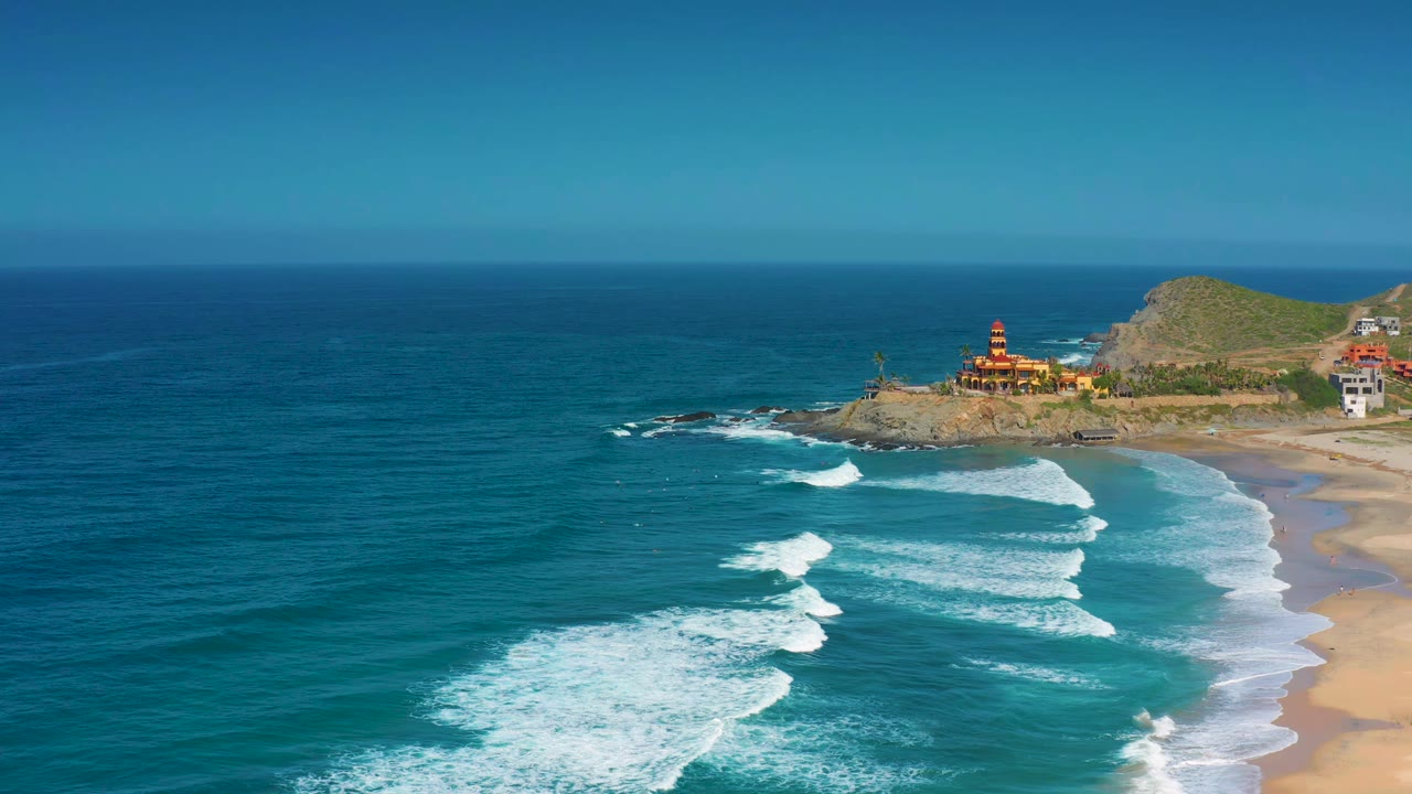 Blue sea and white breaking waves beside a sandy shoreline and coastal buildings.