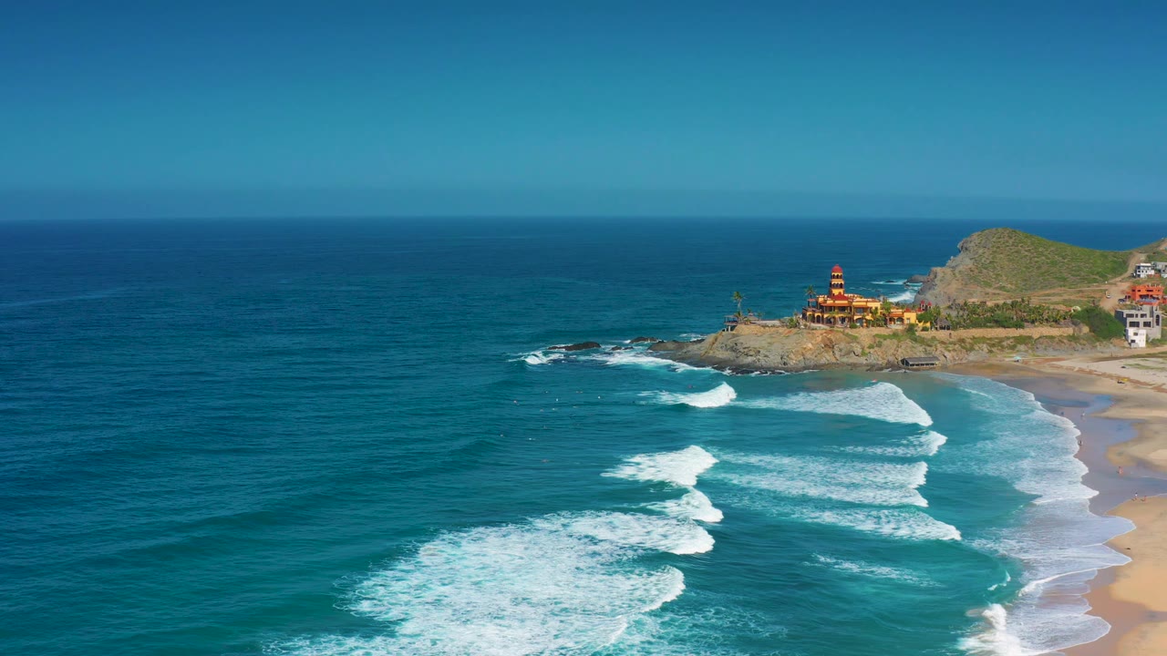 Aerial frame from Cerritos, Baja California Sur showing a broad field of blue ocean, white waves spreading toward the sand, a rocky headland with low buildings, and no visible people.