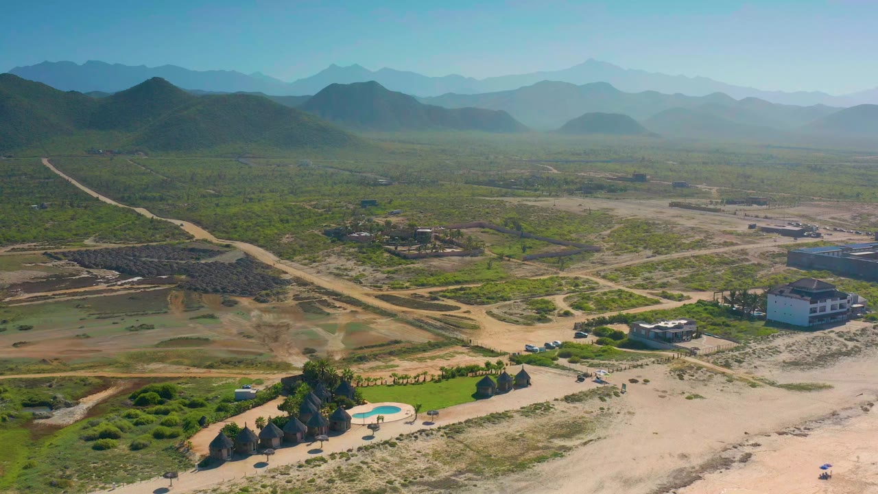 Wide aerial frame over Cerritos, Baja California Sur, with a swimming pool and palm cluster low in frame, scattered homes and dusty roads beyond, and no visible people.