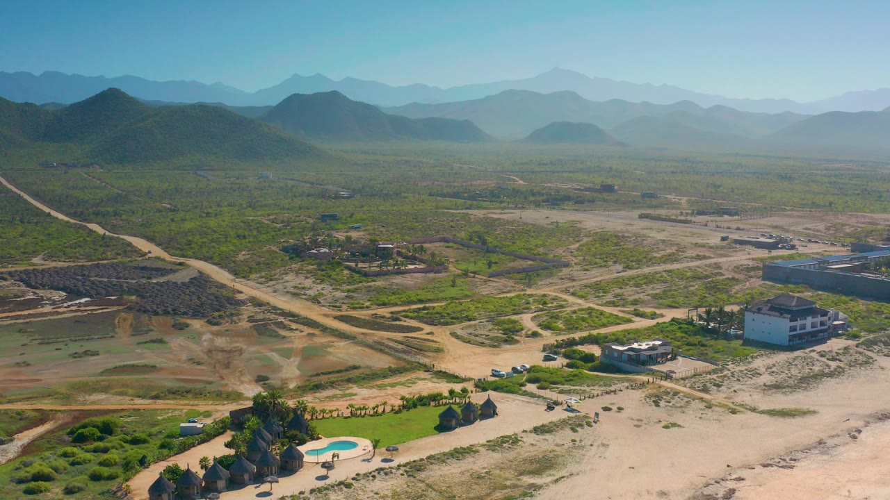 Wide aerial view over Cerritos, Baja California Sur with dry residential plots, scattered houses, a pool area at the lower edge, dirt roads, distant mountains, and no visible people.
