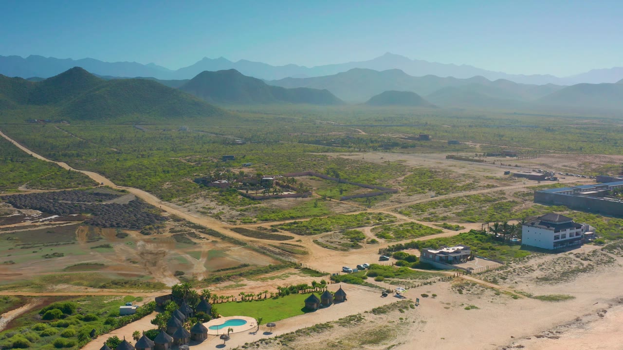 High wide view over Cerritos, Baja California Sur with a small pool near the lower frame, houses, graded dirt tracks, scrubland, layered mountains, and no visible people.