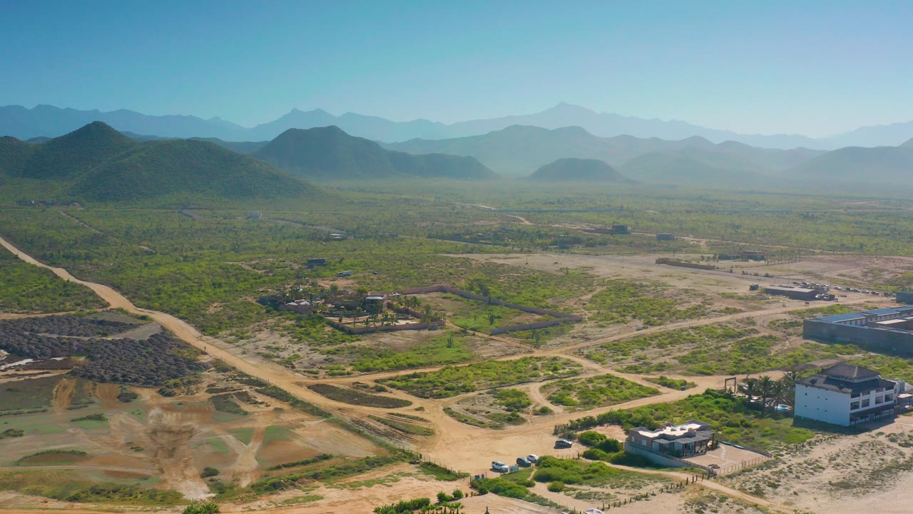 Wide drone view over Cerritos, Baja California Sur, with green scrubland, pale dirt roads, scattered homes, distant hills, blue sky, and no visible people.