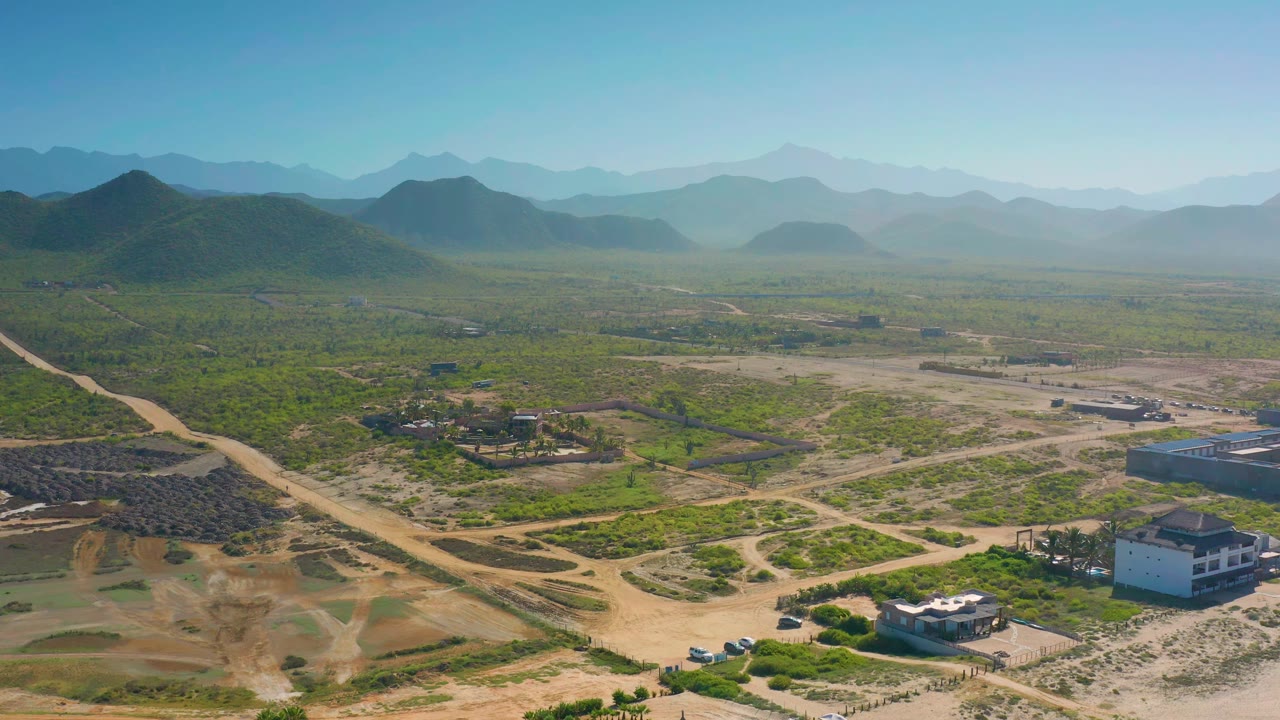 Elevated view across Cerritos, Baja California Sur, showing a central dirt road, patchy green land, several homes, hazy mountain layers, and no visible people.