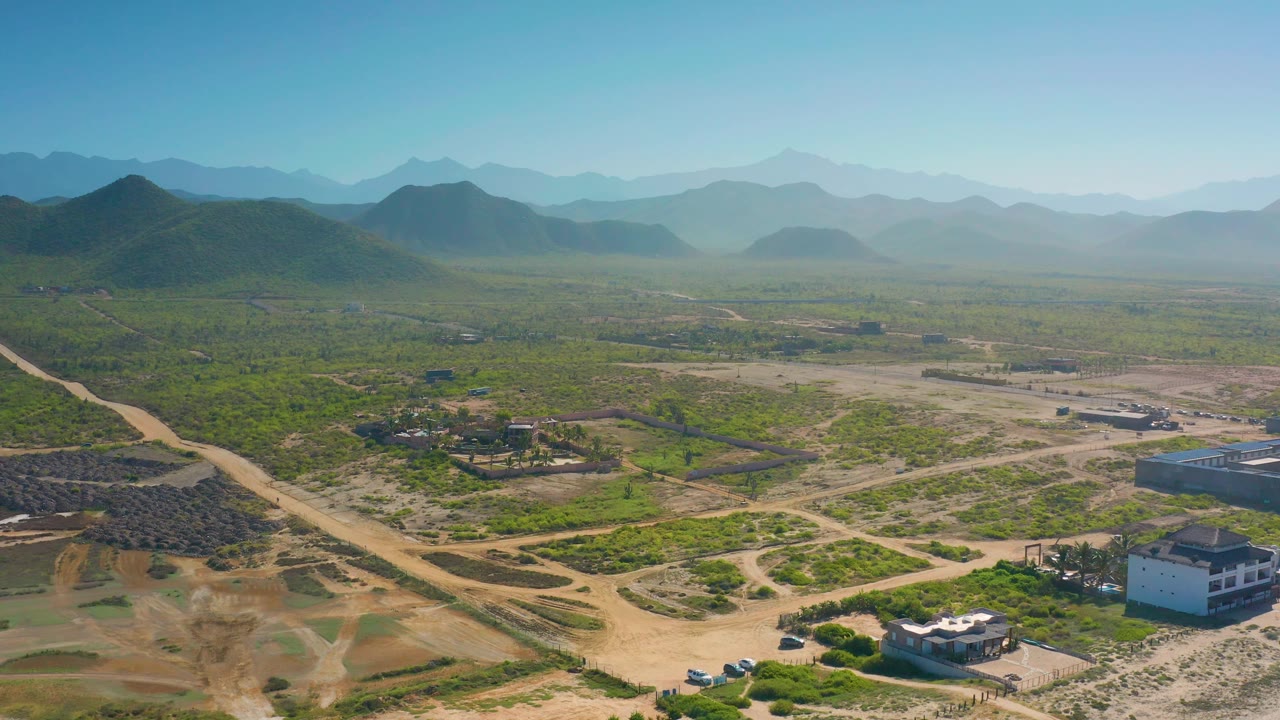 Wide Cerritos aerial with a pale road crossing green terrain, low buildings at the lower edge, layered hills on the horizon, and no visible people.