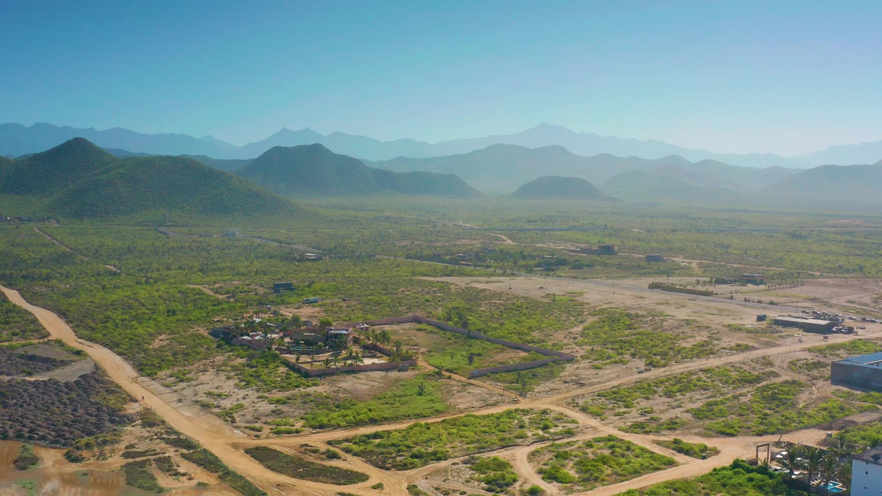 Wide aerial view in Cerritos, Baja California Sur, with dirt roads crossing scrubland, a small building cluster near the lower left, layered hills, hazy mountains, and no visible people.
