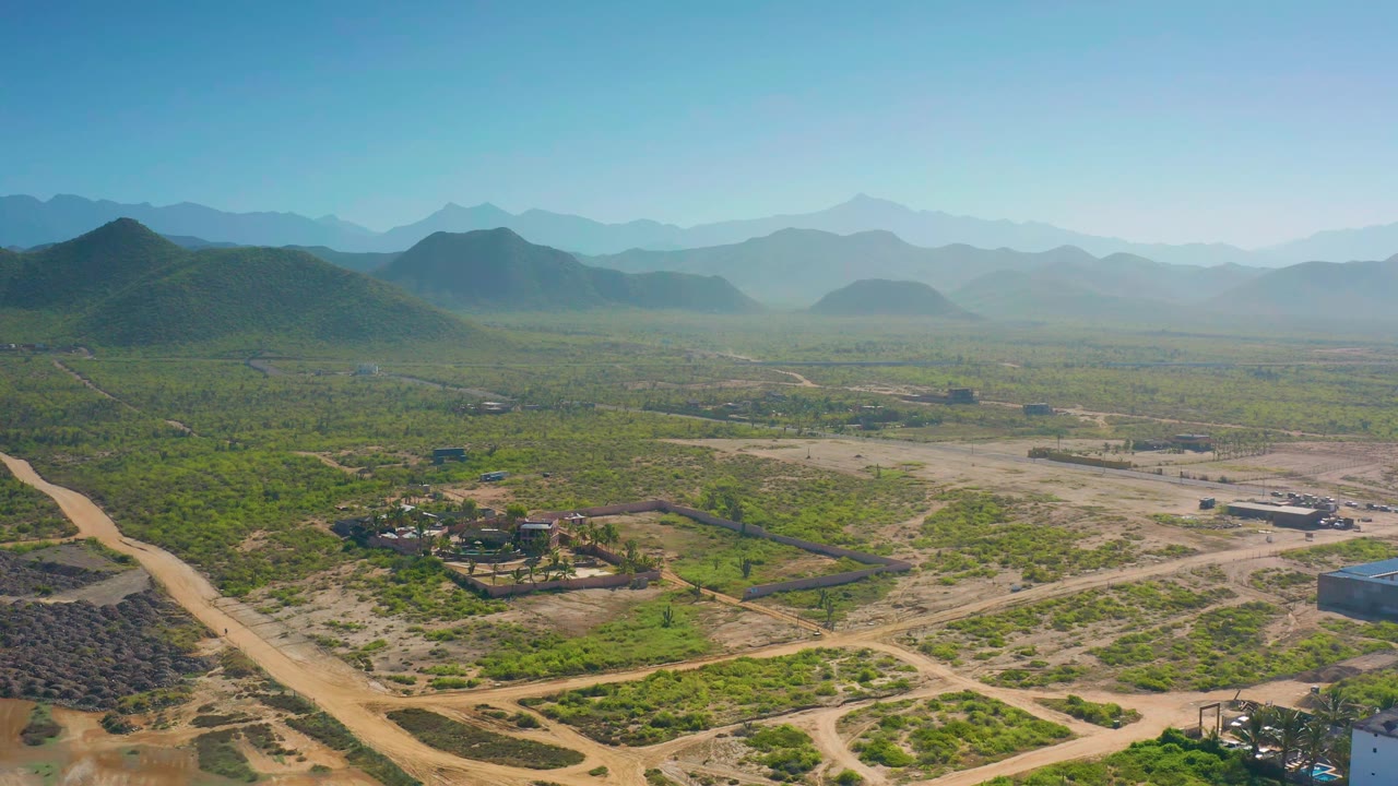 Drone frame over Cerritos countryside showing a dirt road at the lower left, fenced plots and scattered buildings across green scrub, rounded hills beyond, and no visible people.