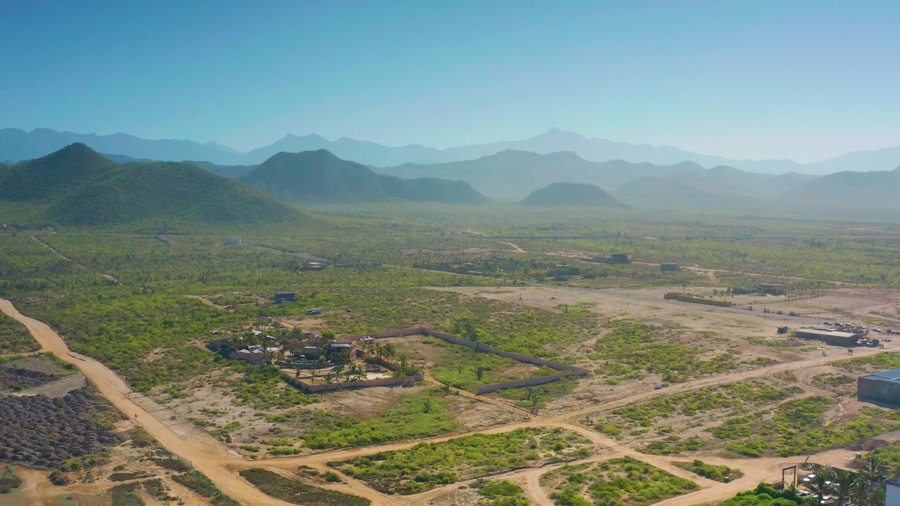 Aerial view in Cerritos, Baja California Sur, with a small compound low in frame, pale dirt tracks across dry scrubland, low hills to the left, a hazy mountain line, and no visible people.