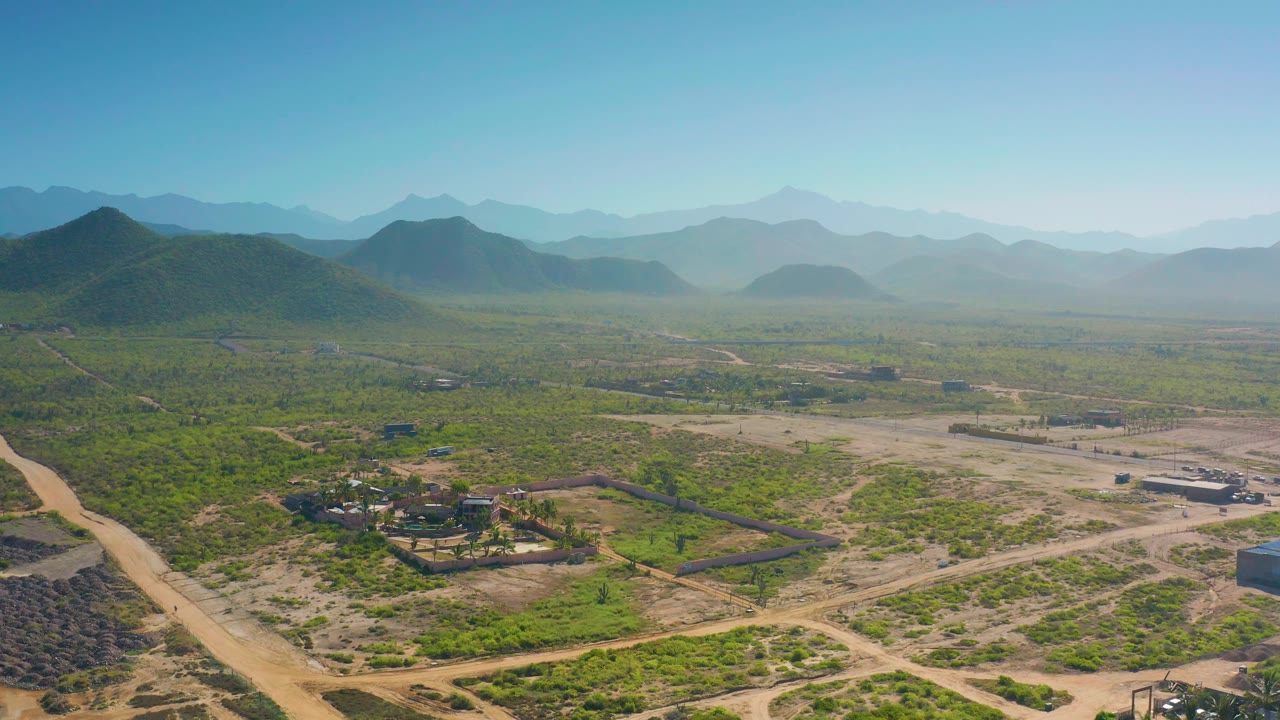 Drone view looking across dirt roads, fenced land, scrub vegetation, hills, and blue mountains.