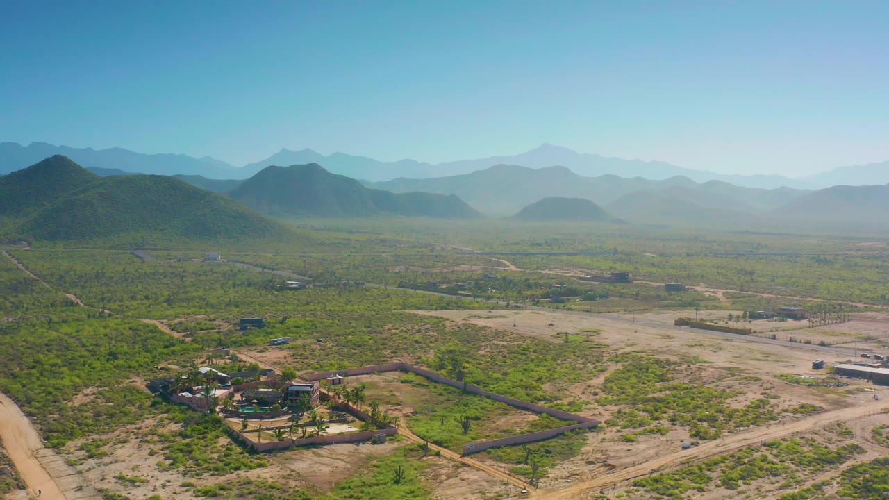 Drone-style landscape frame in Cerritos, Baja California Sur, showing desert lots, a compound near the lower center, scattered structures, dirt tracks, hazy hills, and no visible people.