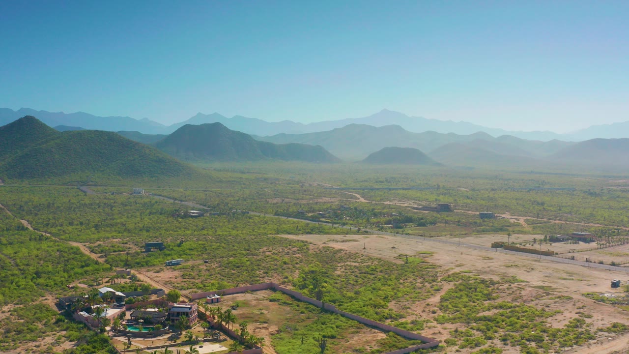 Wide aerial view of scrubland, dirt roads, low buildings, and distant mountains under blue sky.