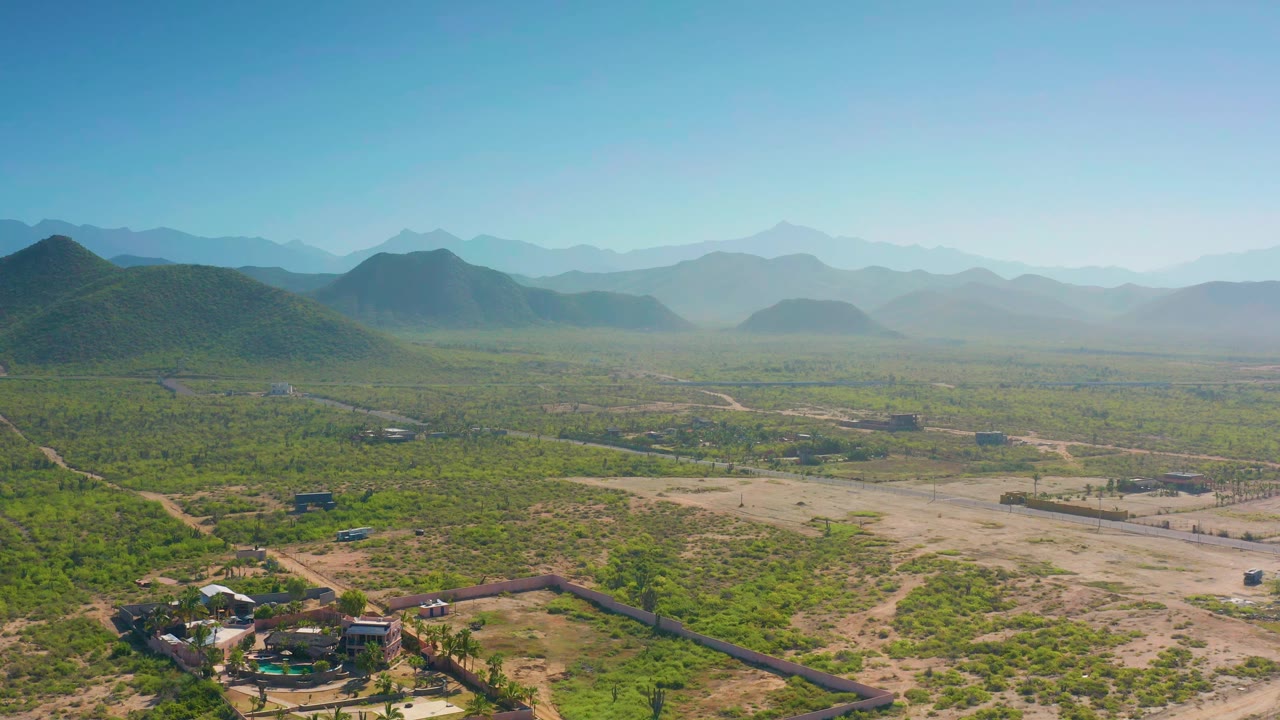 One second into the clip, the Cerritos aerial view keeps the scrubland foreground and scattered structures low in frame, with hazy layered mountains across the horizon and no visible people.