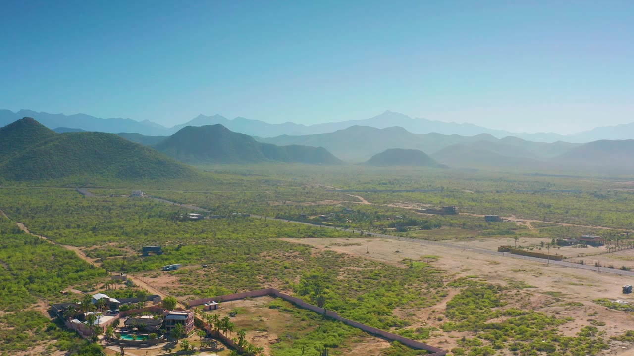 At 0:02, the elevated Cerritos view shows sandy clearings and green scrub in the foreground, a flat plain in the midground, layered mountain ridges in the background, and no visible people.