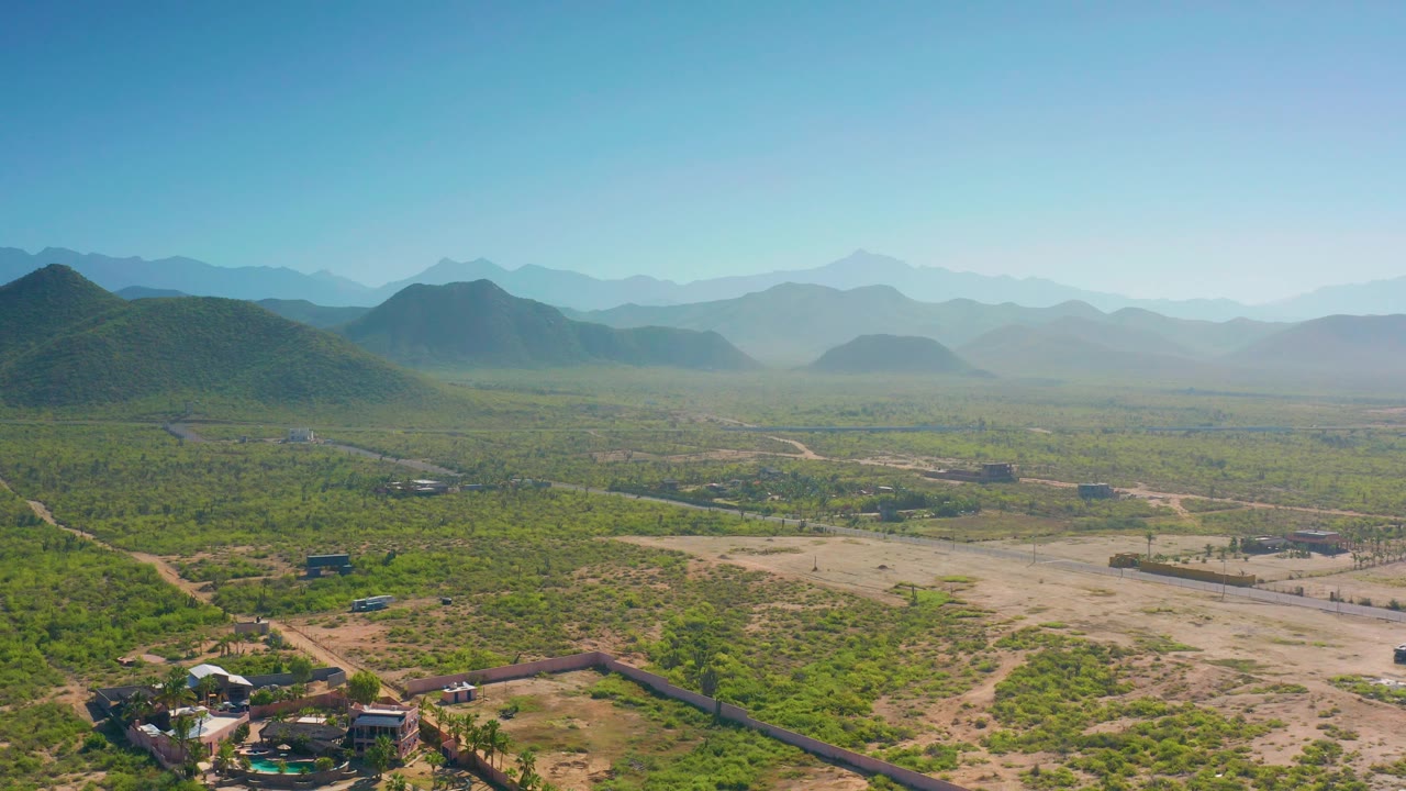 Midway through the clip, the Cerritos landscape remains a wide aerial composition with scrubland, dirt tracks, small buildings near the lower edge, hazy mountain layers, and no visible people.