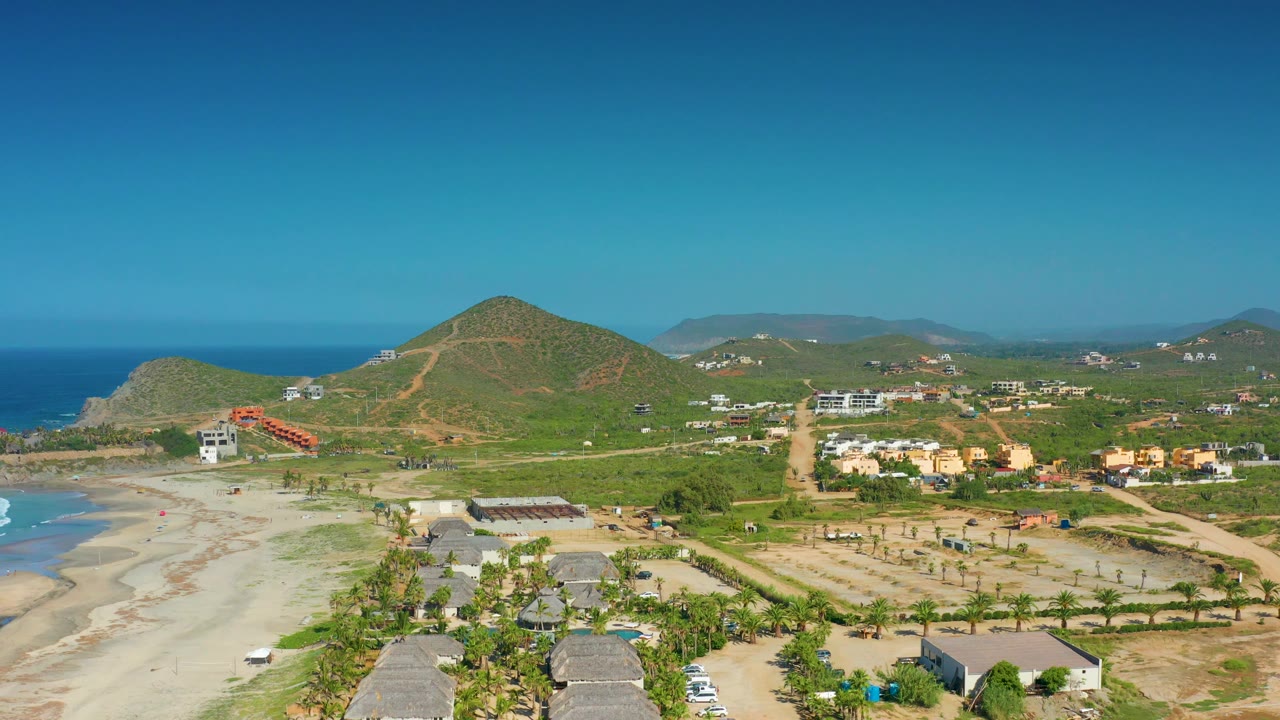 High coastal view of Cerritos, Baja California Sur, showing surf curling at the left edge, sandy flats, palm-lined homes and roads below, scattered buildings inland, rounded green hills, and no visible people.