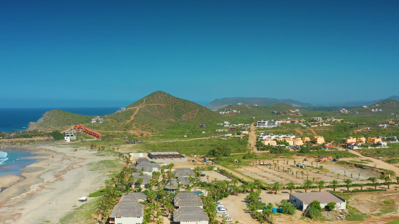 Drone-height view over thatched roofs, beach, roads, and hills