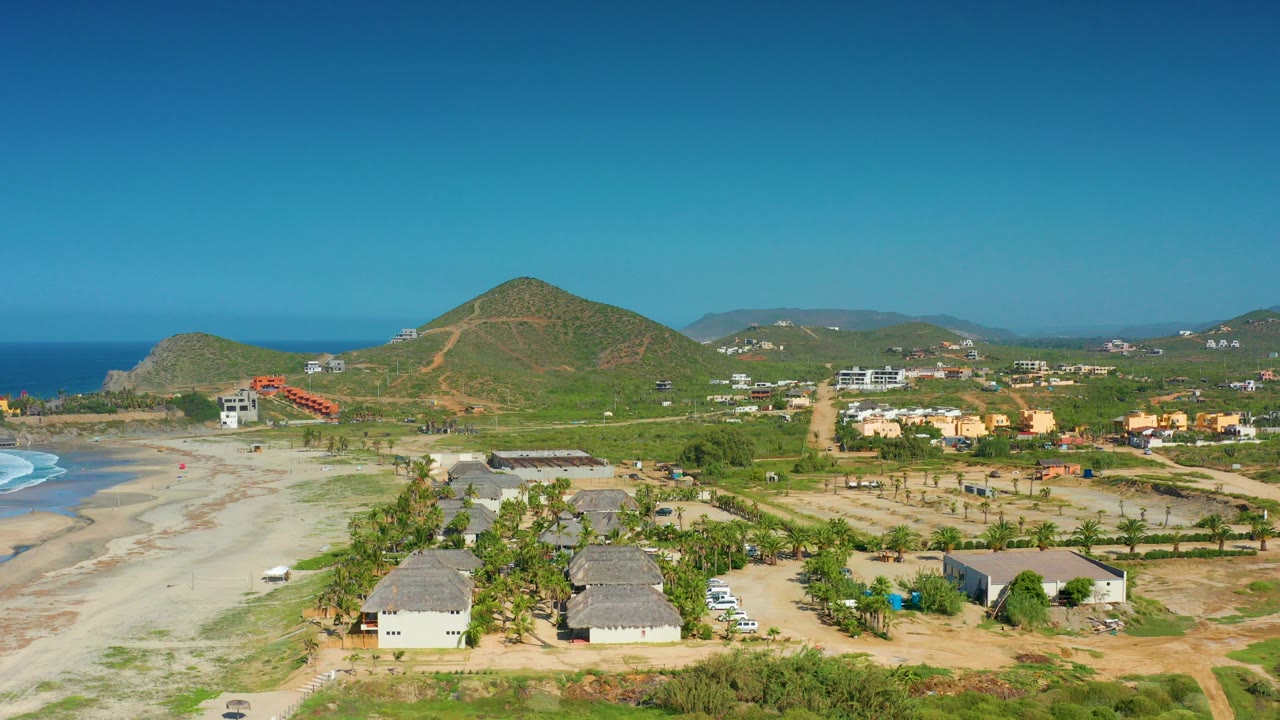 Wide aerial frame over Cerritos, Baja California Sur showing pale beach, white surf, hut-like houses, palms, dirt roads, green hills, blue ocean, and no visible people.