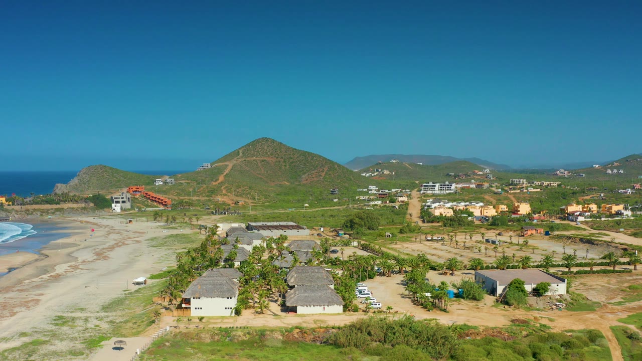A high view of Cerritos beach shows two hut-like structures in the midground, white surf along the sand, scattered homes, green mountains, and no visible people.