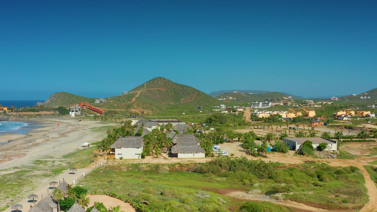 Opening aerial frame over Cerritos, Baja California Sur, with the beach on the left, low houses and dirt tracks below, hills in the distance, and no visible people.