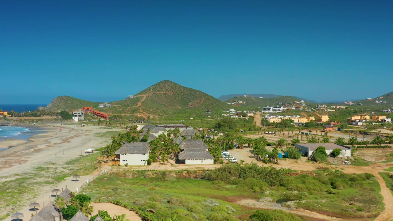 Second aerial frame of Cerritos showing a wider spread of sandy shoreline at left, clustered low buildings near the center, green scrub in front, and no visible people.