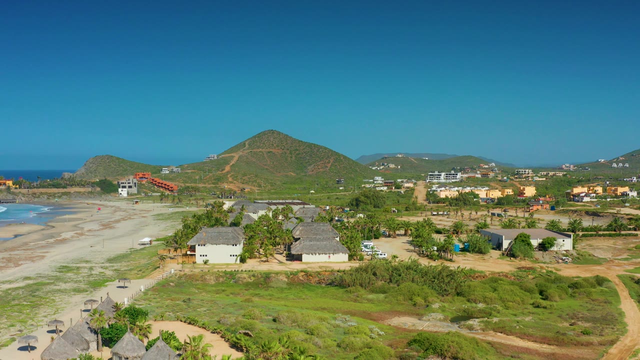 Third frame over Cerritos with thatched roofs near the lower edge, a pale beach running along the left side, scattered houses inland, and no visible people.