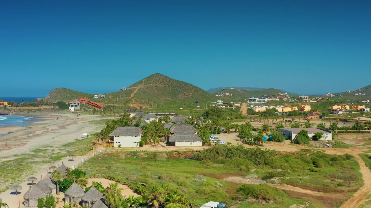 Aerial town-and-beach view with foreground thatched roofs, pool, houses, and hills.