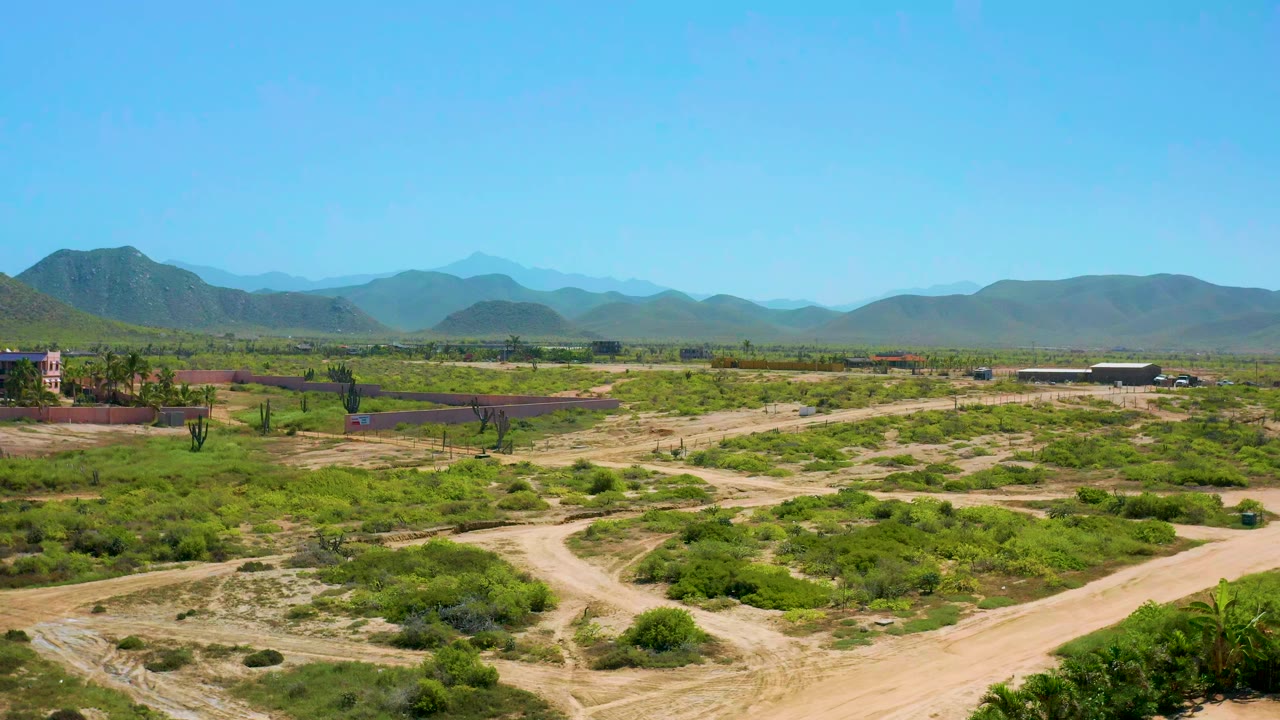 Opening aerial frame over Cerritos, Baja California Sur with tan dirt roads in the foreground, low green desert scrub, a small compound at left, layered mountains beyond, and no visible people.