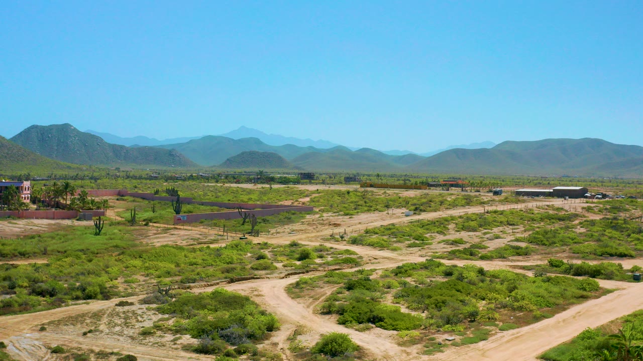 Early aerial frame over Cerritos, Baja California Sur, with pale dirt roads in the foreground, green scrubland across the middle distance, blue mountains on the horizon, and no visible people.