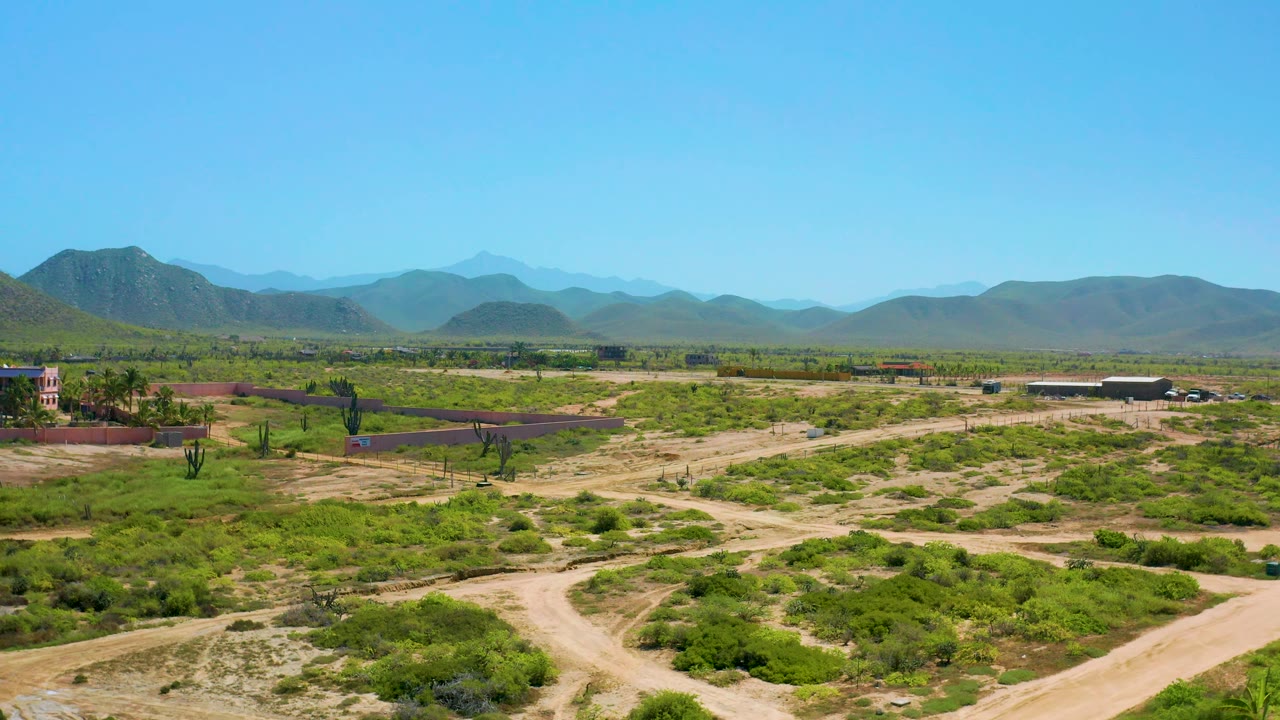 Cerritos, Baja California Sur appears from above with open scrubland, winding dirt paths, scattered cacti, a small building at frame edge, hazy mountains, and no visible people.