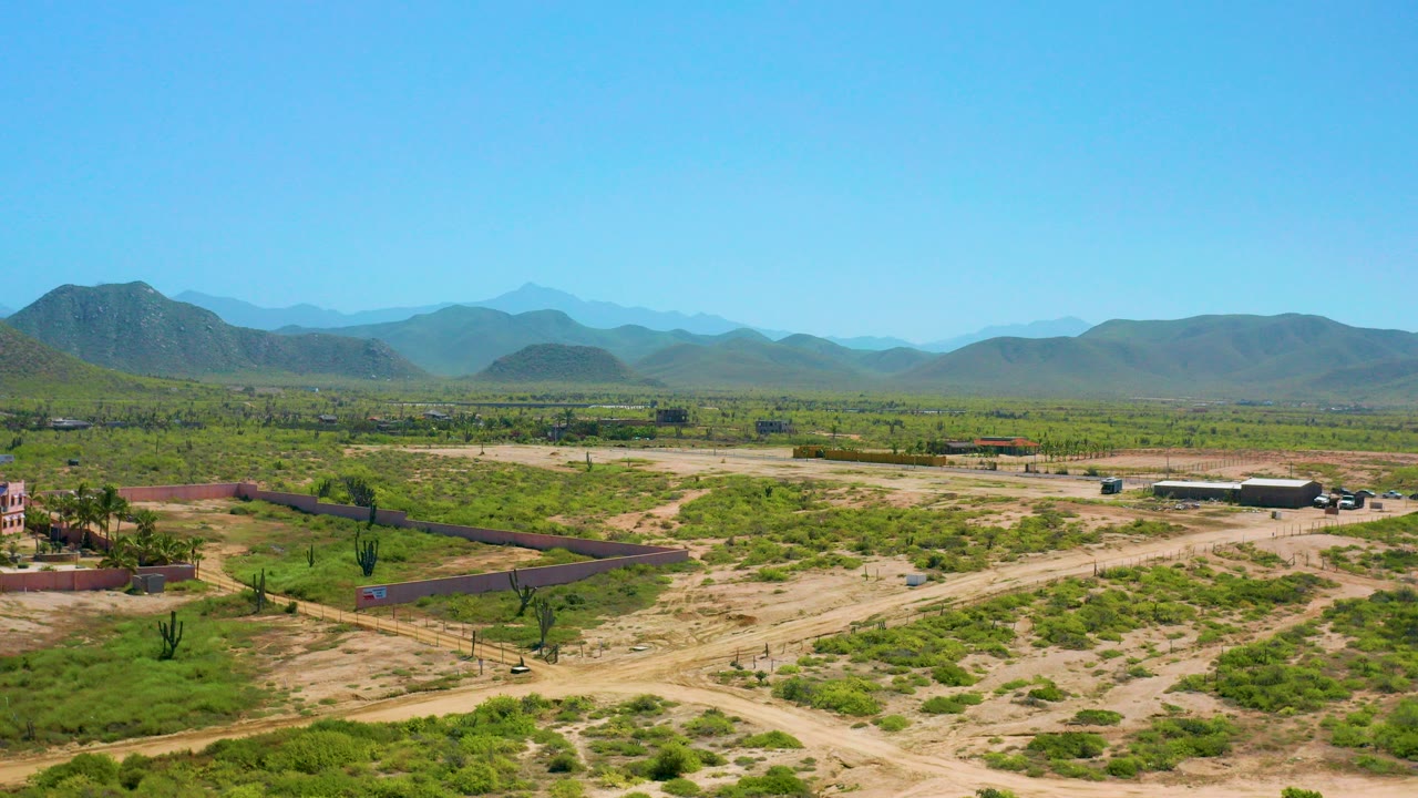 Wide drone frame over Cerritos, Baja California Sur with sandy roads, green scrub, low property walls, scattered buildings, distant mountain ridges, and no visible people.