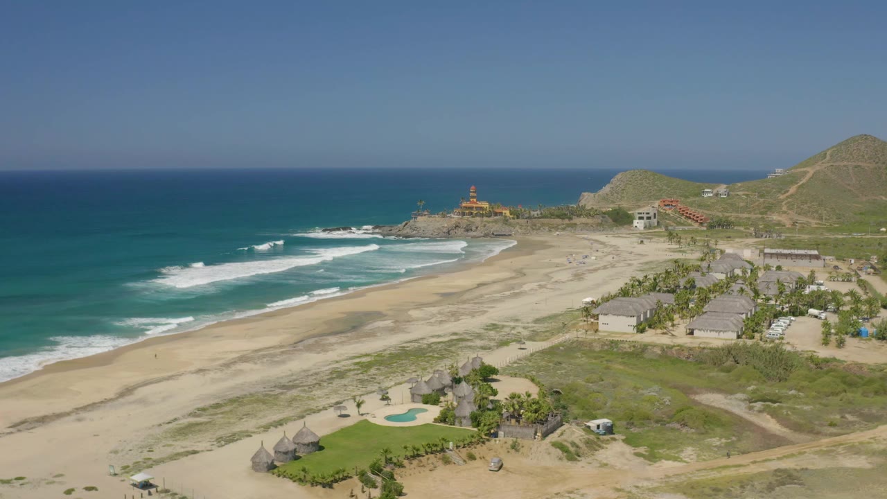 Aerial frame from Cerritos, Baja California Sur with turquoise water, breaking waves, a broad sandy beach, palapa-style roofs near a small pool, hillside roads, and no visible people.