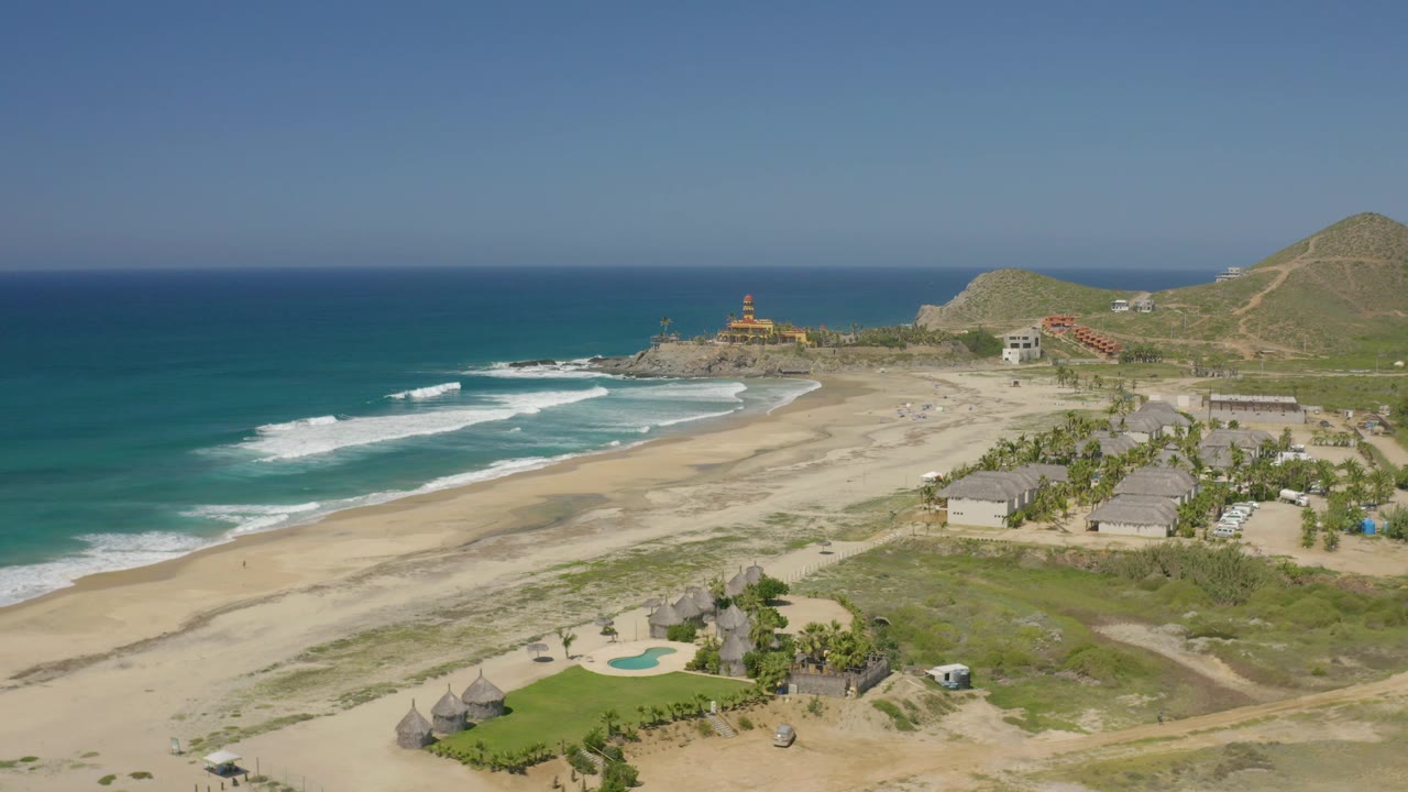 Cerritos aerial view showing white surf bands rolling toward an open sandy beach, scrub-covered foreground, low buildings, hillside tracks, and no visible people.