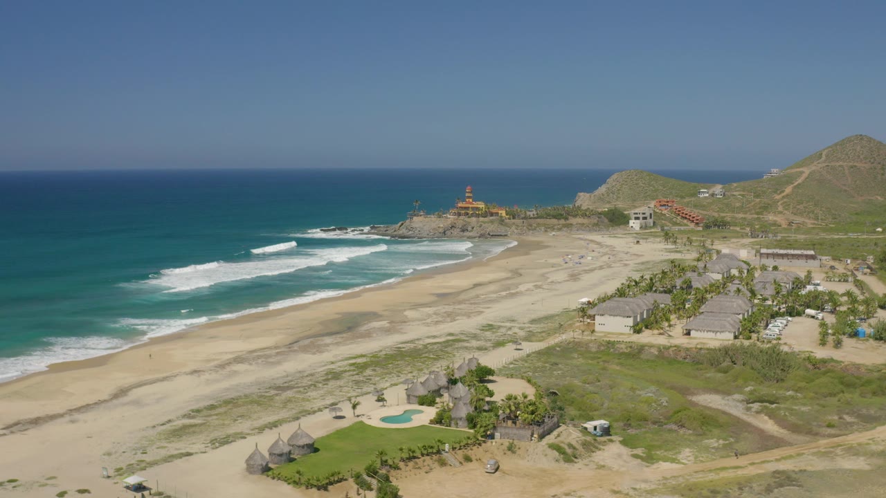 Wide beach aerial with ocean, surf, resort roofs, and hillside.