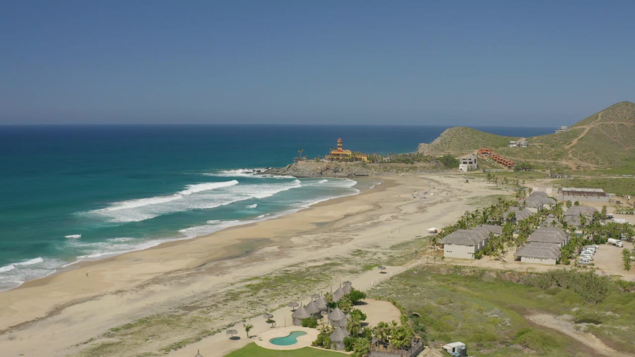 Wide beach aerial with waves, shoreline buildings, palms, hills, and open sky.