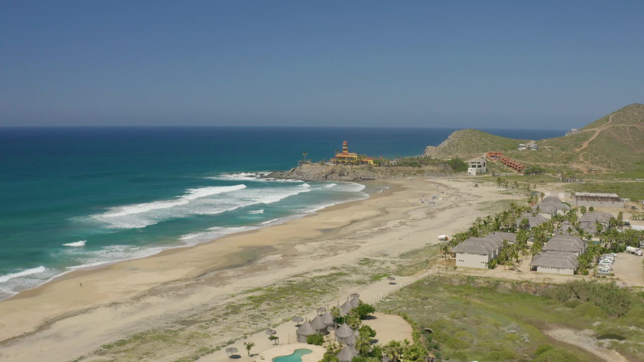 Aerial frame over Cerritos, Baja California Sur, with the ocean filling the left side, surf bands near shore, beach structures at right, hills beyond, and no visible people.