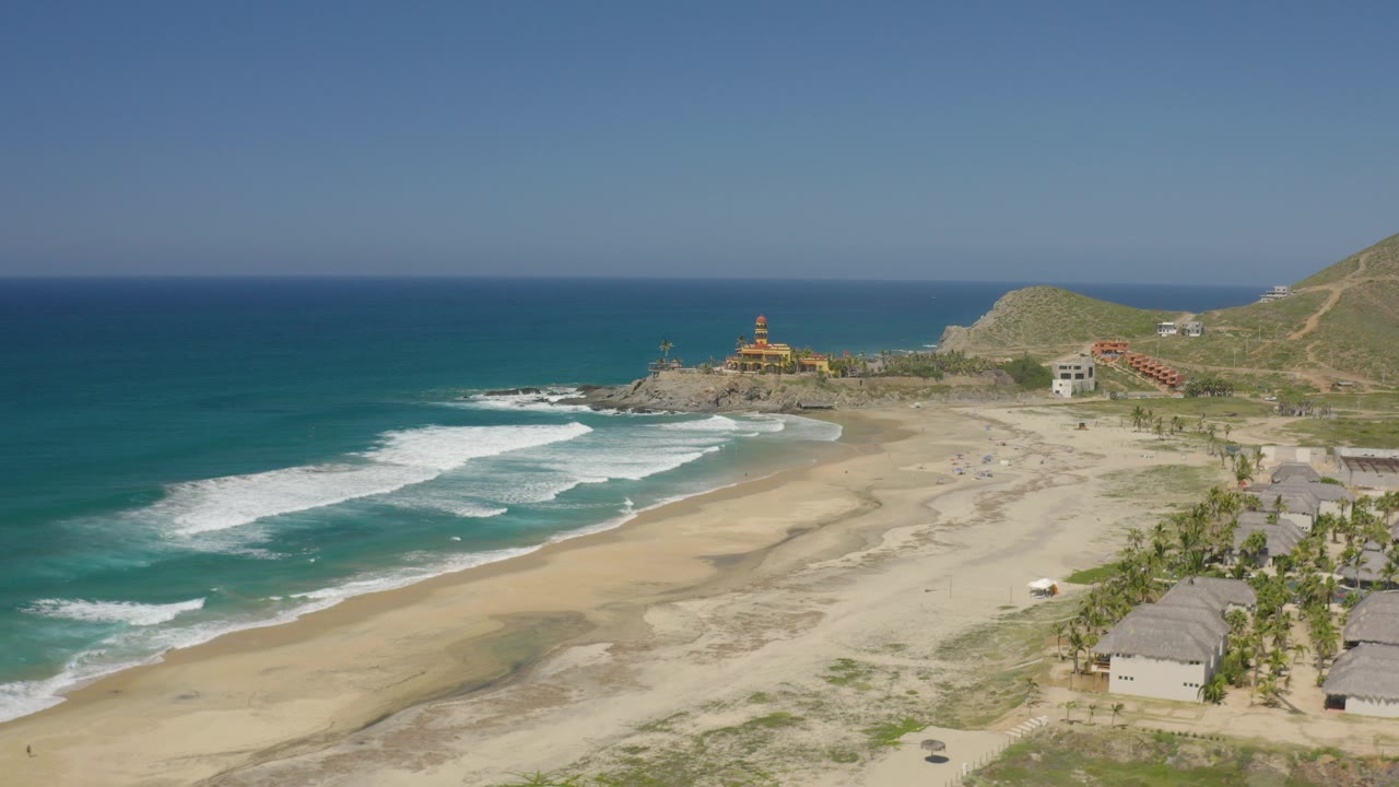 Opening aerial frame over Cerritos, Baja California Sur, showing blue water, white surf, wide sand, beach huts, palms, hillside roads, and no visible people.