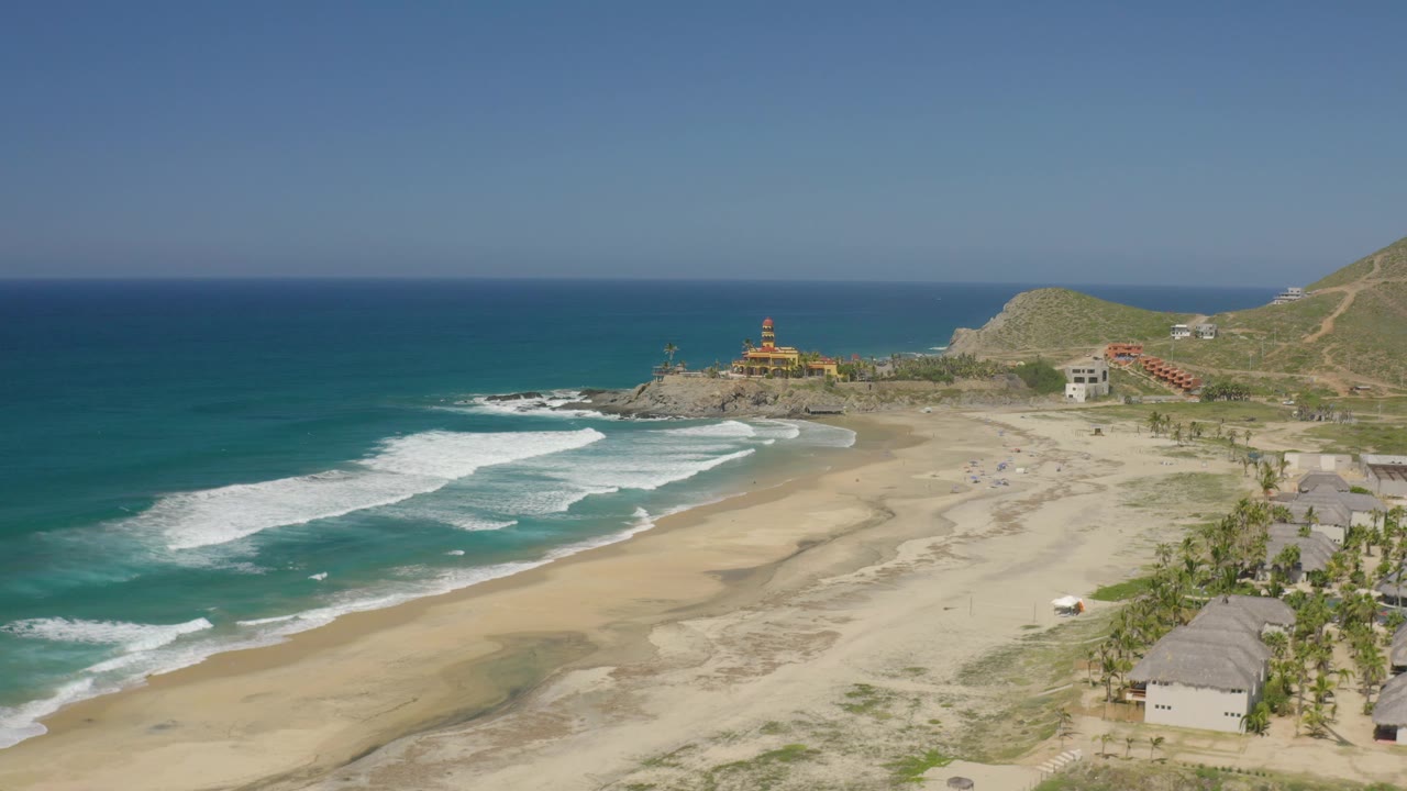 A high coastal view of Cerritos, Baja California Sur, with foamy waves in the left foreground, dry and wet sand across the center, right-side huts and palms, and no visible people.