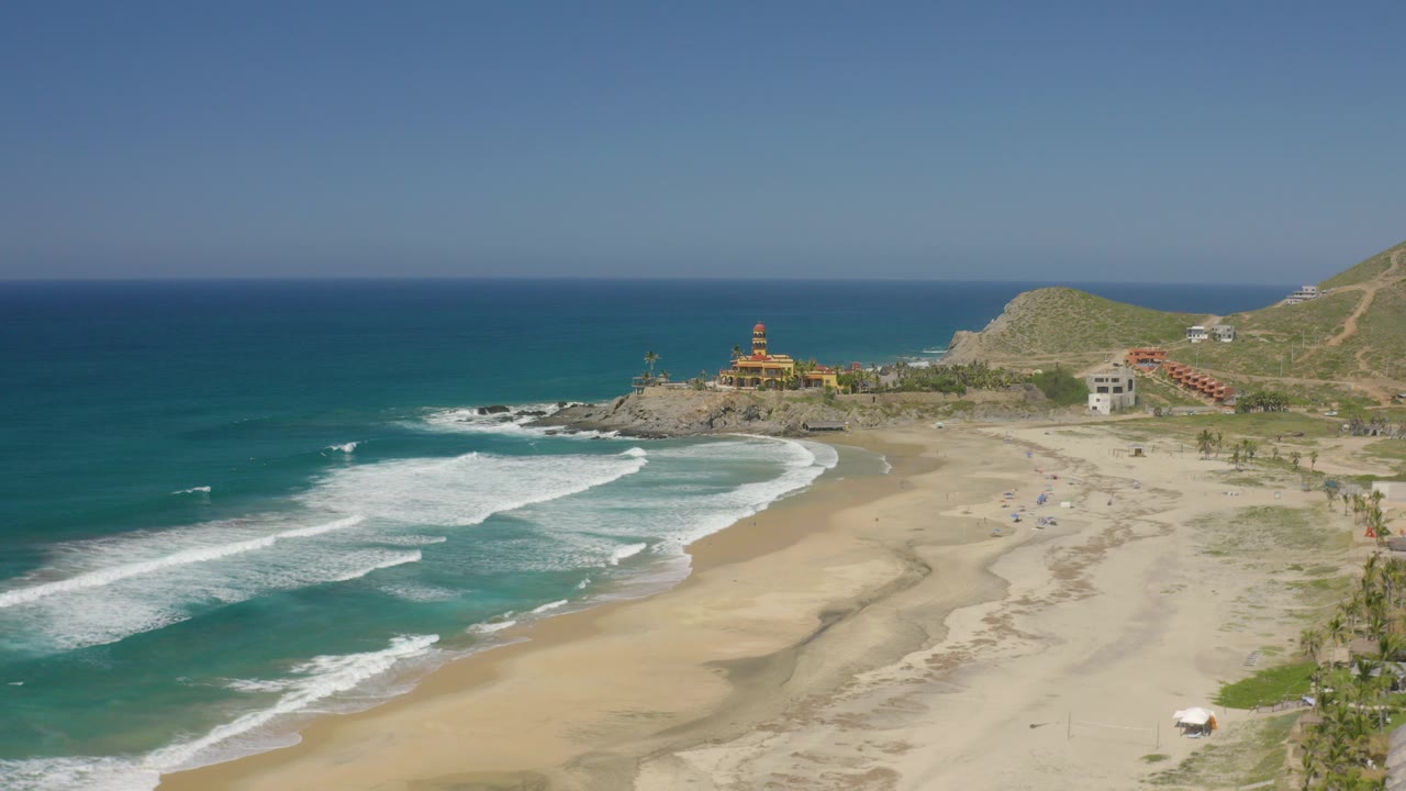 Aerial frame over Cerritos, Baja California Sur showing blue water, white surf bands, a wide sandy beach, low hills, coastal buildings, and no visible people.