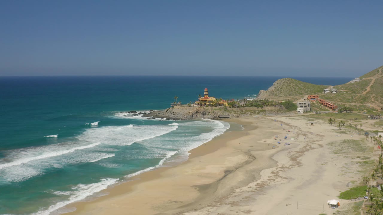 From above Cerritos, Baja California Sur, the frame shows turquoise water, white wave foam, broad sand, a rocky point with structures, dry hills, and no visible people.